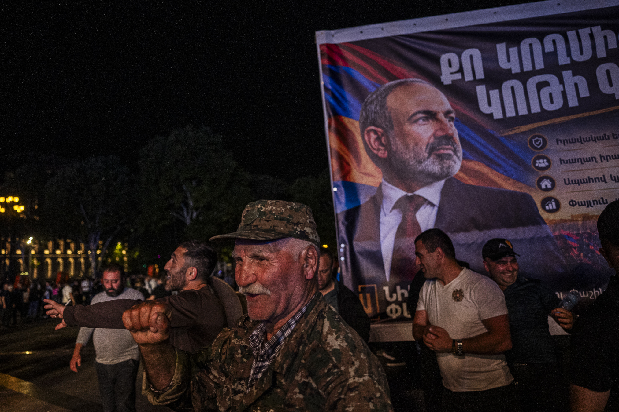 A veteran of the First Nagorno-Karabakh War at a rally of Nikol Pashinyan's party. Behind him is a portrait of Pashinyan from the Revolution. Despite the loss of Karabakh, he supports the current prime minister because 