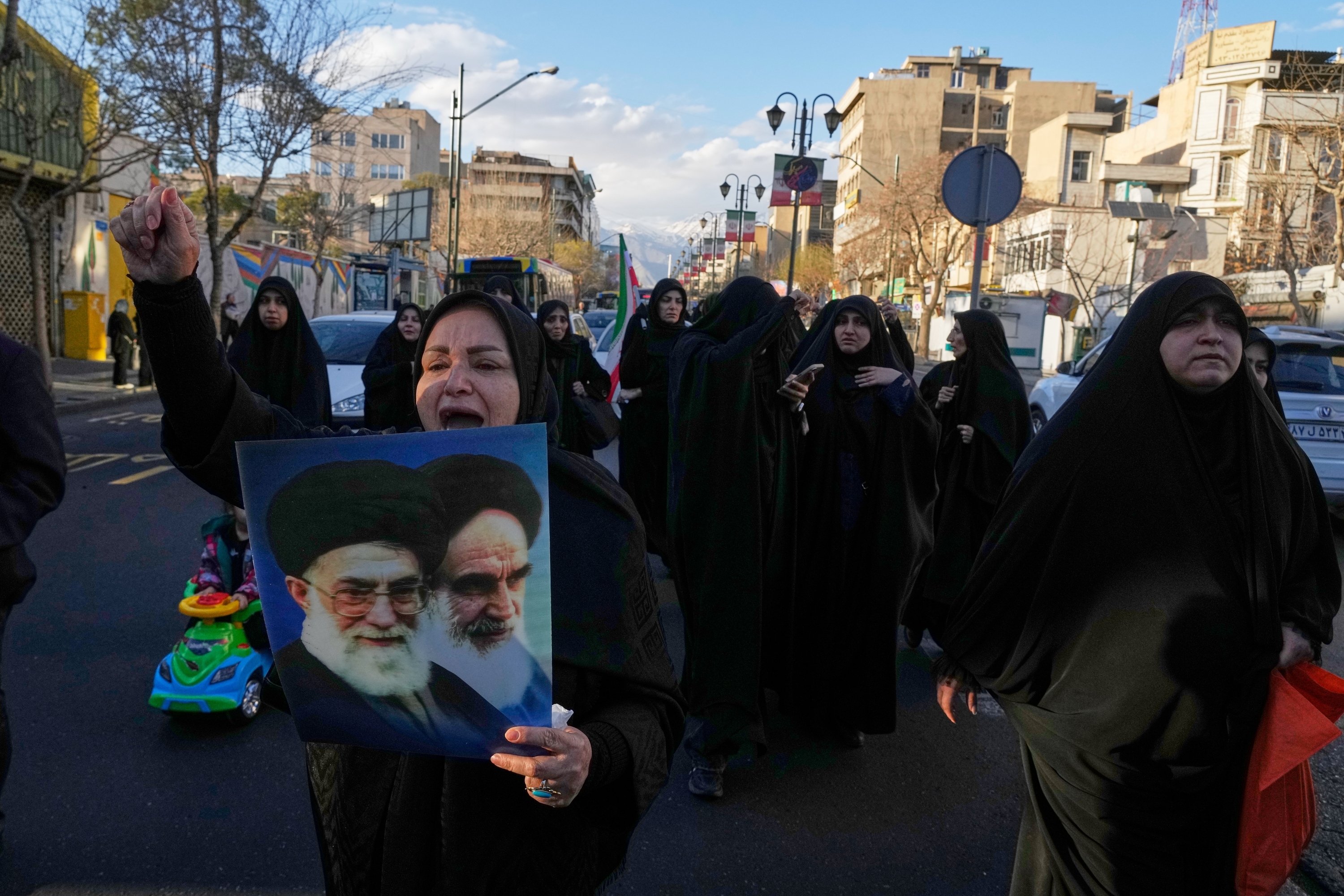 A woman holds pictures of Ayatollah Khomeini and the late Iranian Supreme Leader Ayatollah Ali Khamenei as a group of government supporters march toward Khamenei's residency in Tehran, Iran, Sunday, March 1, 2026, following the confirmed death of Khamenei in U.S. and Israeli strikes. ©AP Photo/Vahid Salemi