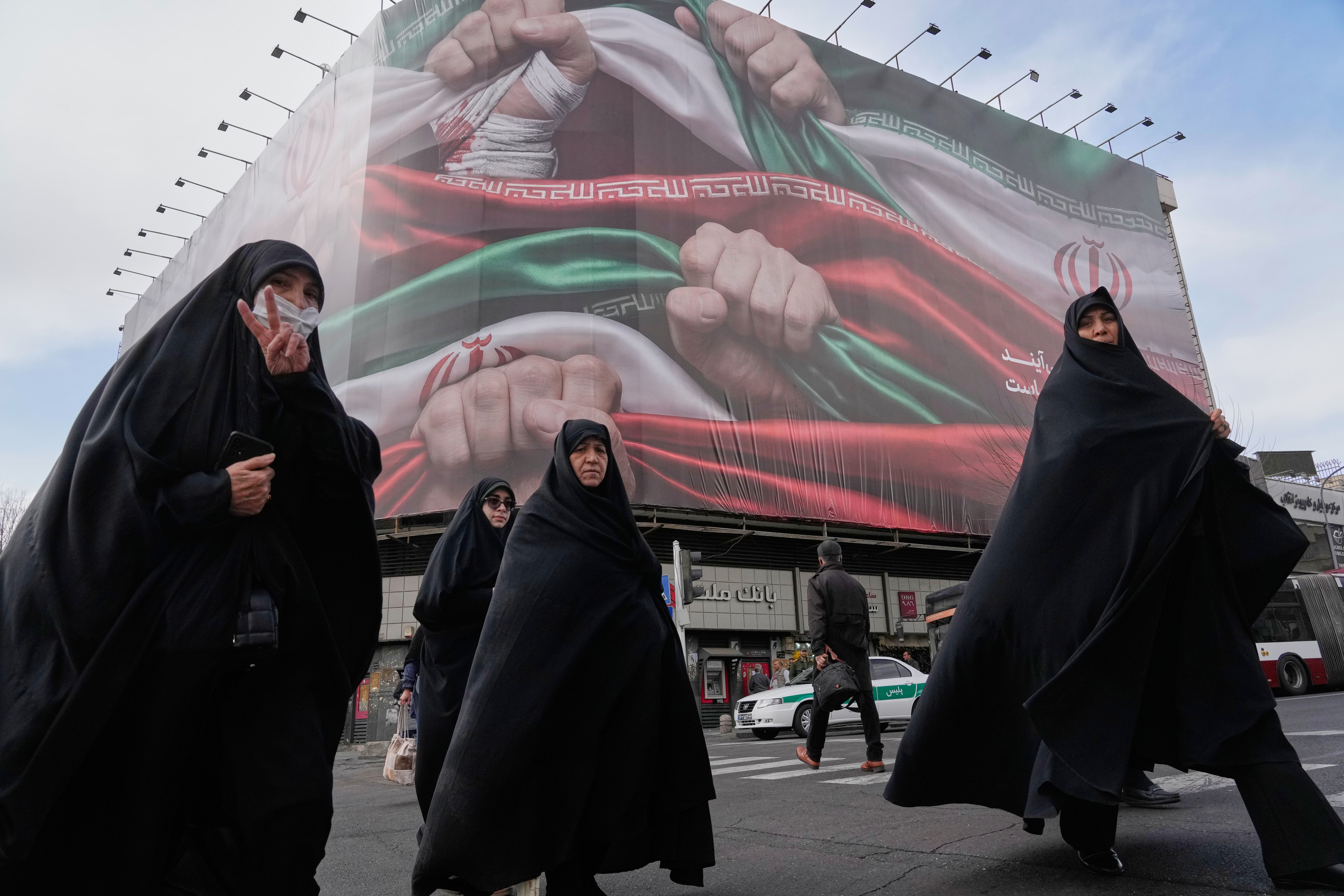 Women, one flashing a victory hand gesture, cross a street under a huge banner showing hands firmly holding Iranian national flags as a sign of patriotism, in Tehran, Iran, Jan. 14, 2026. ©AP Photo/Vahid Salemi